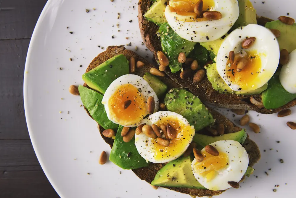 Deilig avocadotoast med bløtkokte egg og pinjekjerner på en hvit tallerken.
