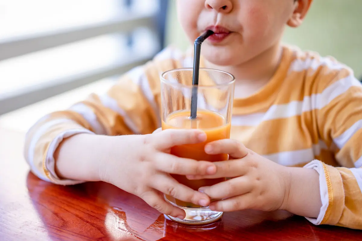 Multivitamin til barn: Nærbilde av et lite barn som drikker fersk appelsinjuice gjennom et sugerør innendørs, symboliserer sunn ernæring og naturlige vitaminer for barn.