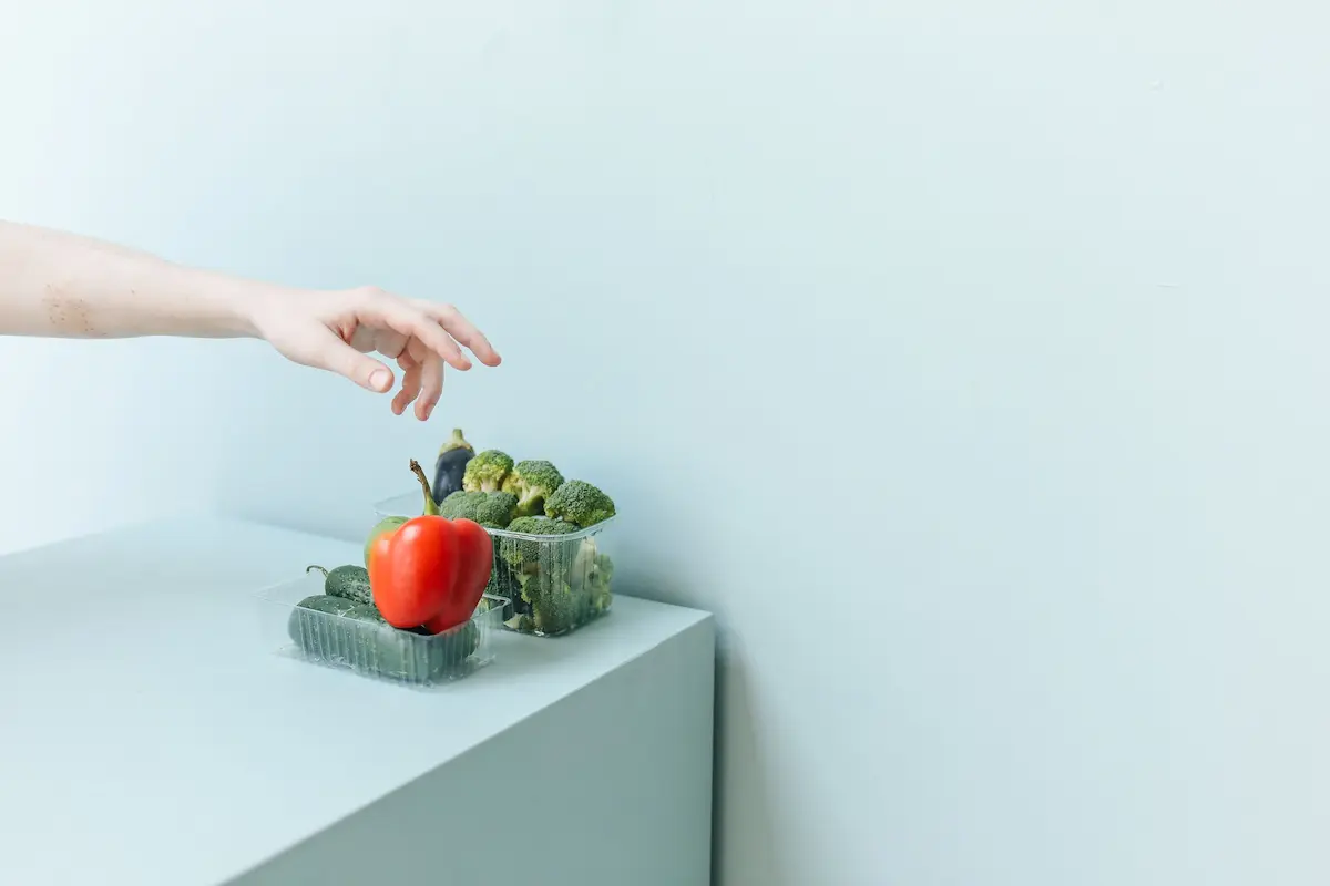 Hånd som rekker etter ferske grønnsaker på en minimalistisk hvit overflate.