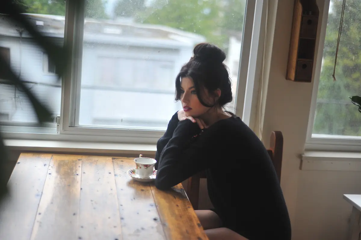 Kvinne som sitter ved et vindu med en kaffekopp, i rolig og ettertenksom stilling innendørs